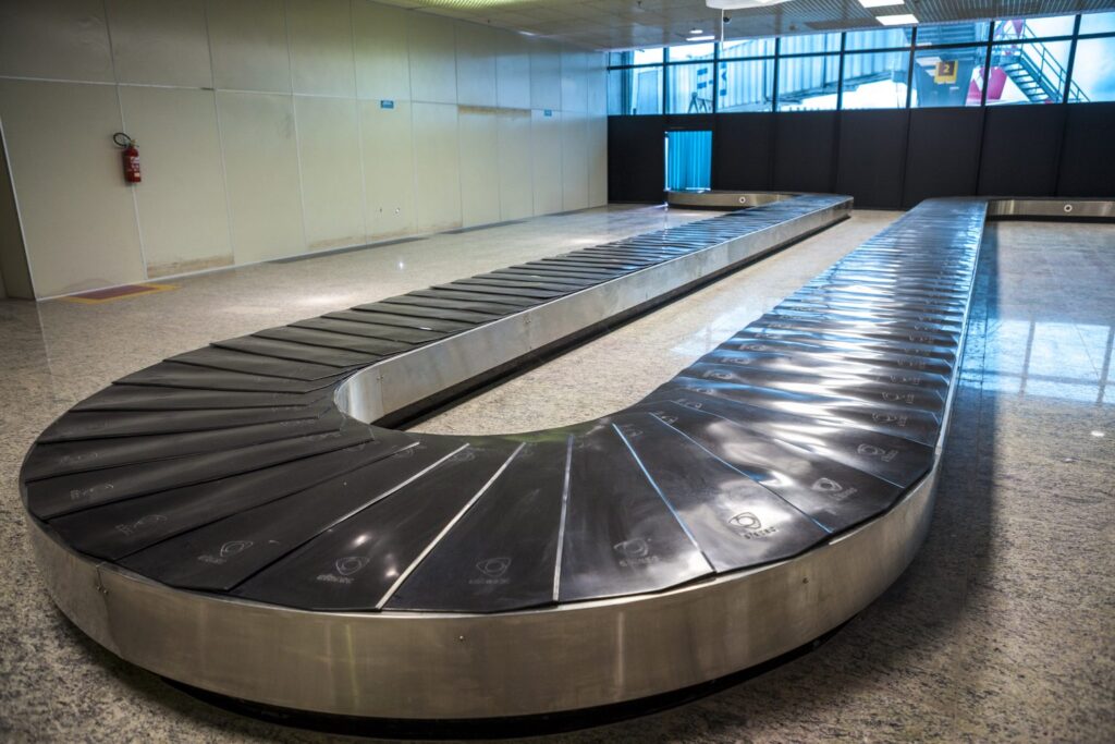 Esteira de aeroporto vazia, na sala não tem ninguém, apenas a esteira, paredes brancas e chão claro. A saida da esteira tem parede preta com janelas em cima.