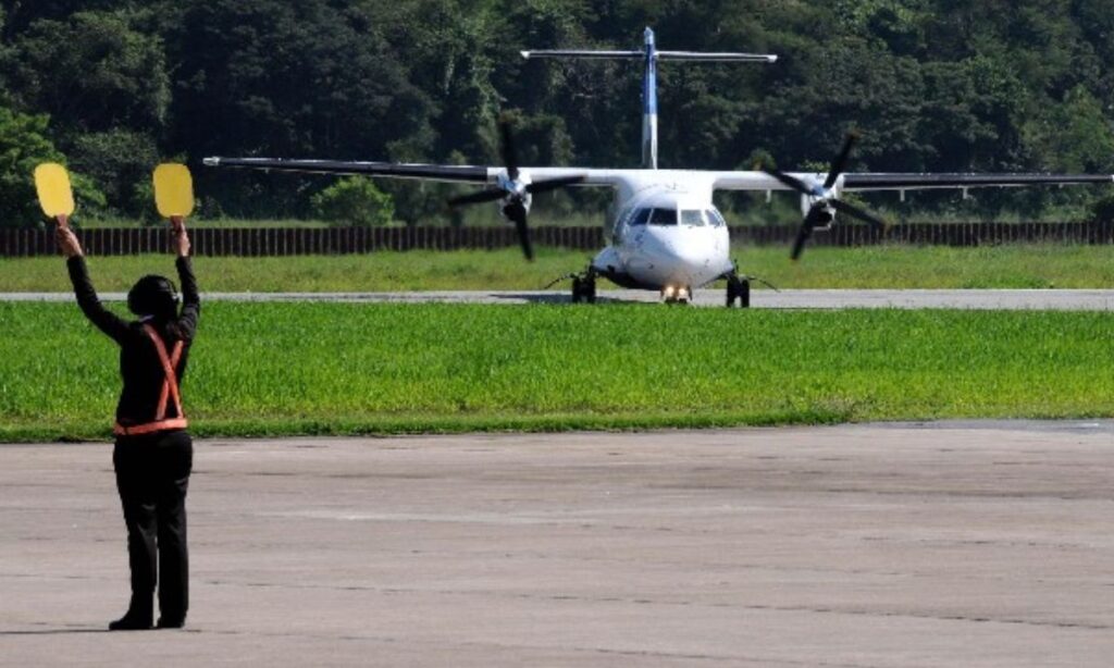 Avião ao fundo manobrando na pista de pouso, no primeiro plano um manobrista de avião fazendo sinal.
